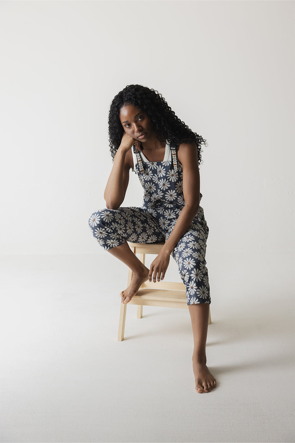 Woman wearing navy overalls with white daisy pattern, sitting on wooden stool