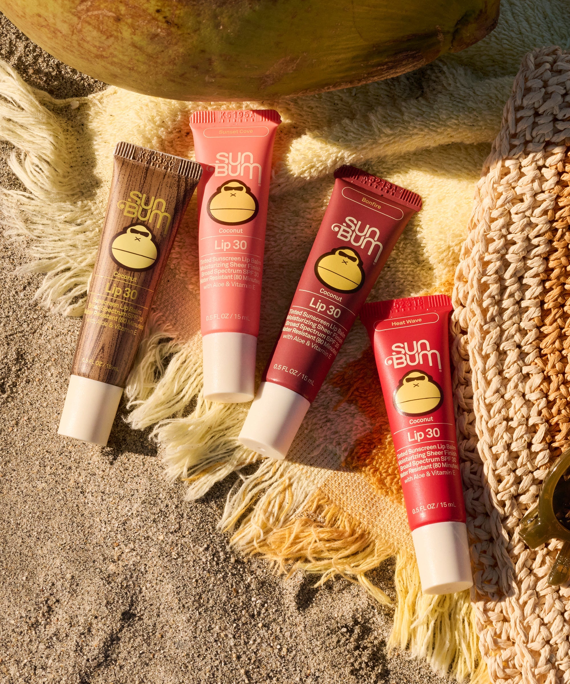 Four tinted lip balm tubes in various shades arranged on sandy beach setting with rope accessories