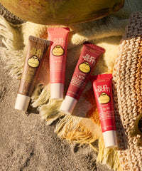 Four tinted lip balm tubes in various shades arranged on sandy beach setting with rope accessories