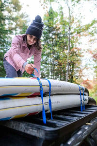 Person securing kayaks to roof rack using blue cam straps with NRS branding visible