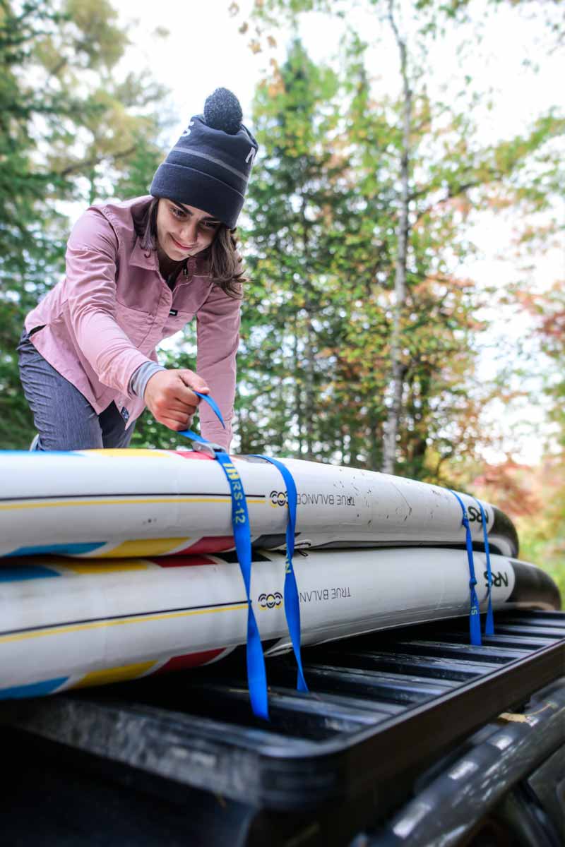 Person securing kayaks to roof rack using blue cam straps with NRS branding visible