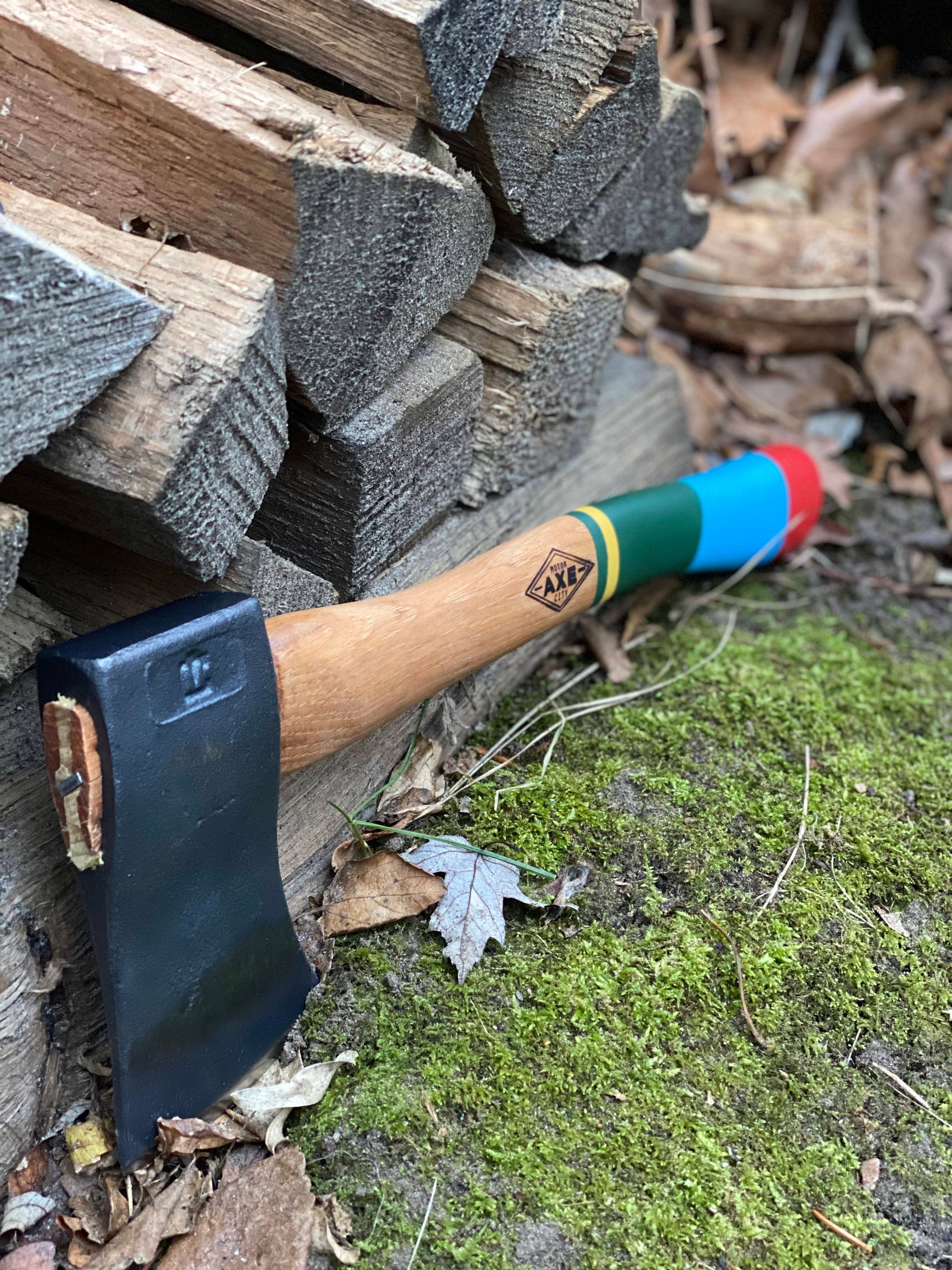 Small camping hatchet with colorful striped wooden handle lying on mossy ground near split wood