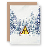 Greeting card showing watercolor winter scene with triangular cabin surrounded by snow-covered evergreen trees
