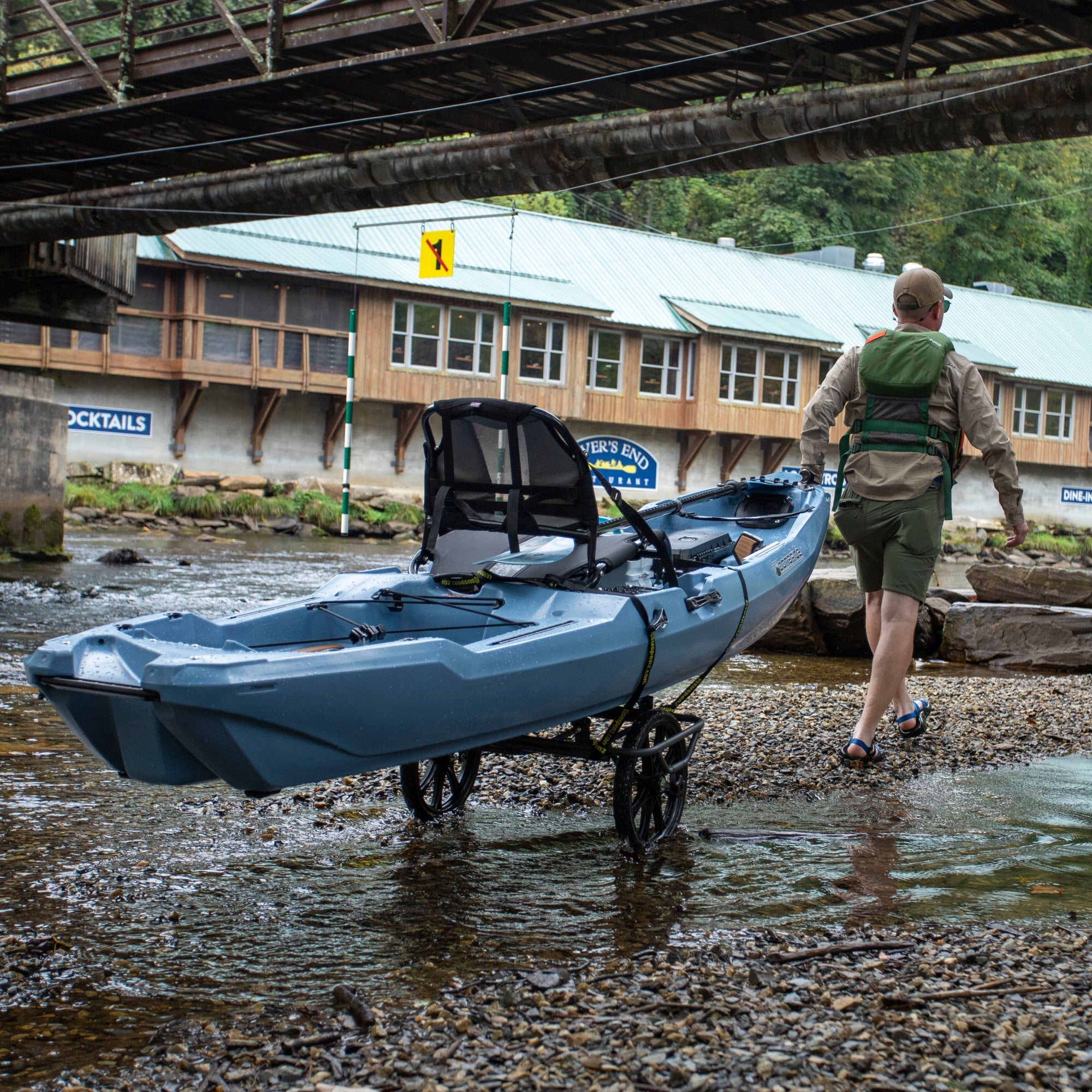 Black wheeled cart carrying blue kayak being pulled by person near water and bridge