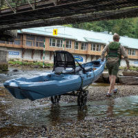 Black wheeled cart carrying blue kayak being pulled by person near water and bridge