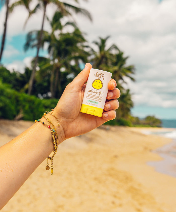 Hand holding small white and yellow mineral sunscreen stick on tropical beach background