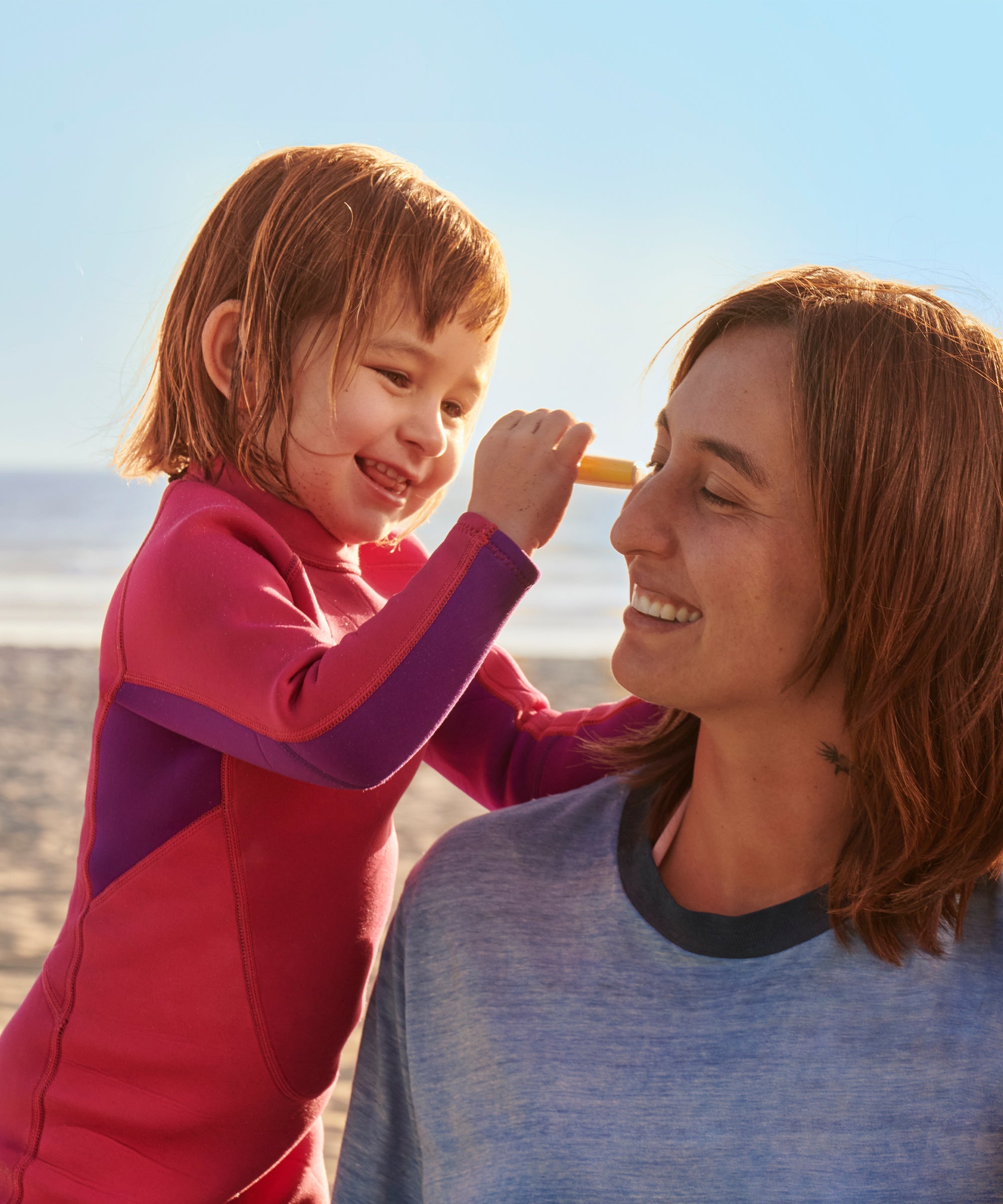 Child applying orange mineral sunscreen stick to woman's nose at beach