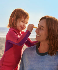 Child applying orange mineral sunscreen stick to woman's nose at beach
