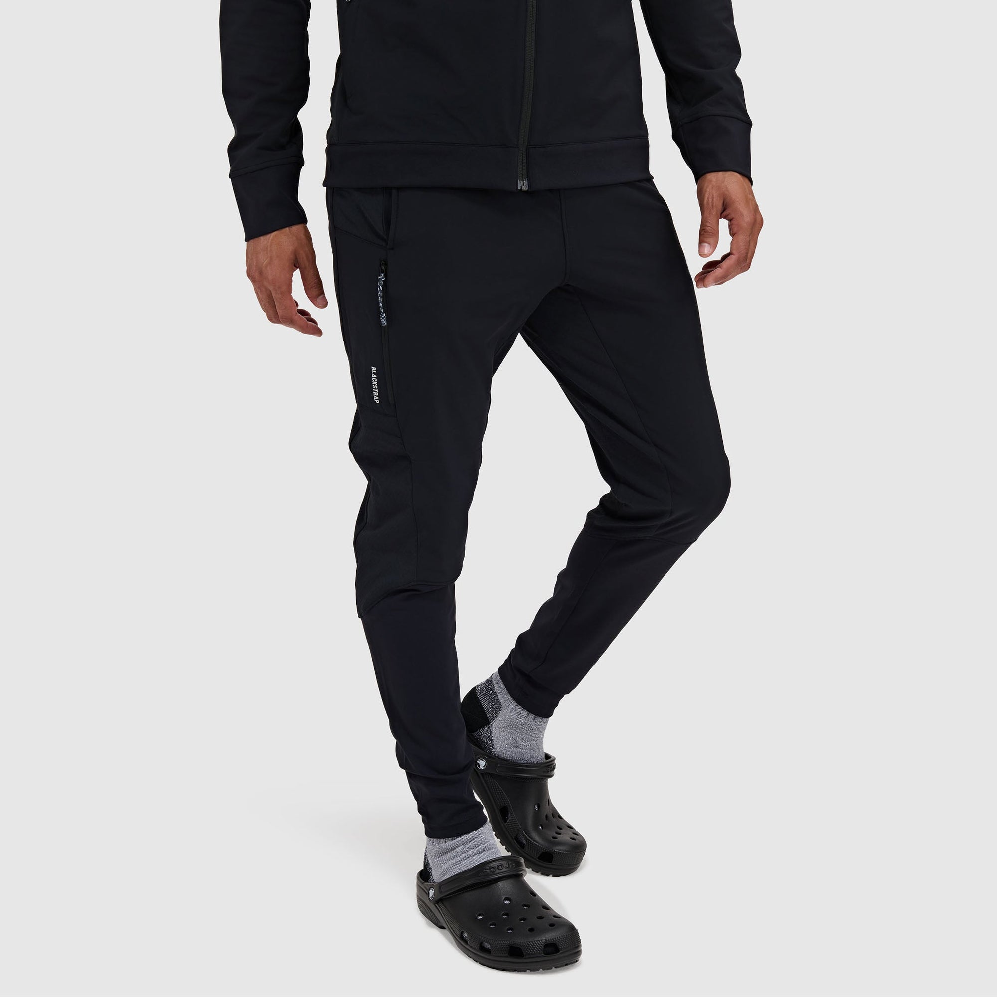 Men's slim-fit black jogger pants with zippered side pockets, worn with crocs, front angle view