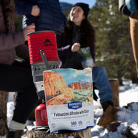 Freeze-dried meal pouch with desert landscape artwork and portable camping stove in snowy outdoor setting