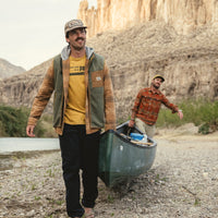 Man carrying a canoe wearing a green fleece vest over a plaid shirt outdoors