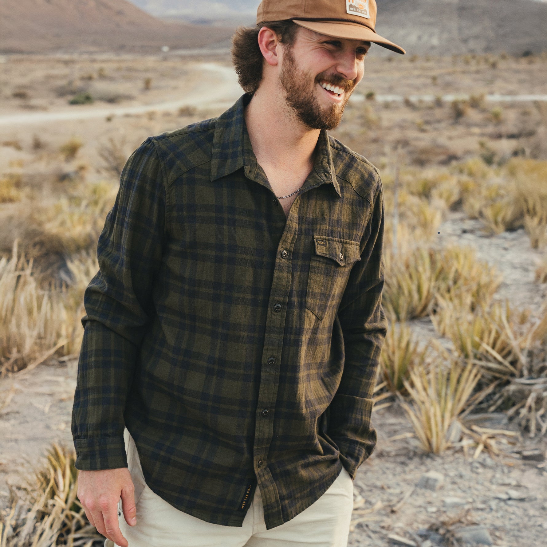 Man wearing olive plaid flannel shirt with snap buttons in desert landscape, smiling