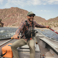 Man rowing a drift boat on a river wearing a plaid flannel shirt, smiling outdoors