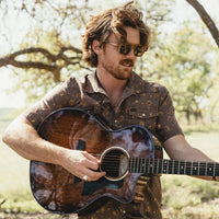 Man playing acoustic guitar outdoors wearing a dark brown floral snap shirt with short sleeves