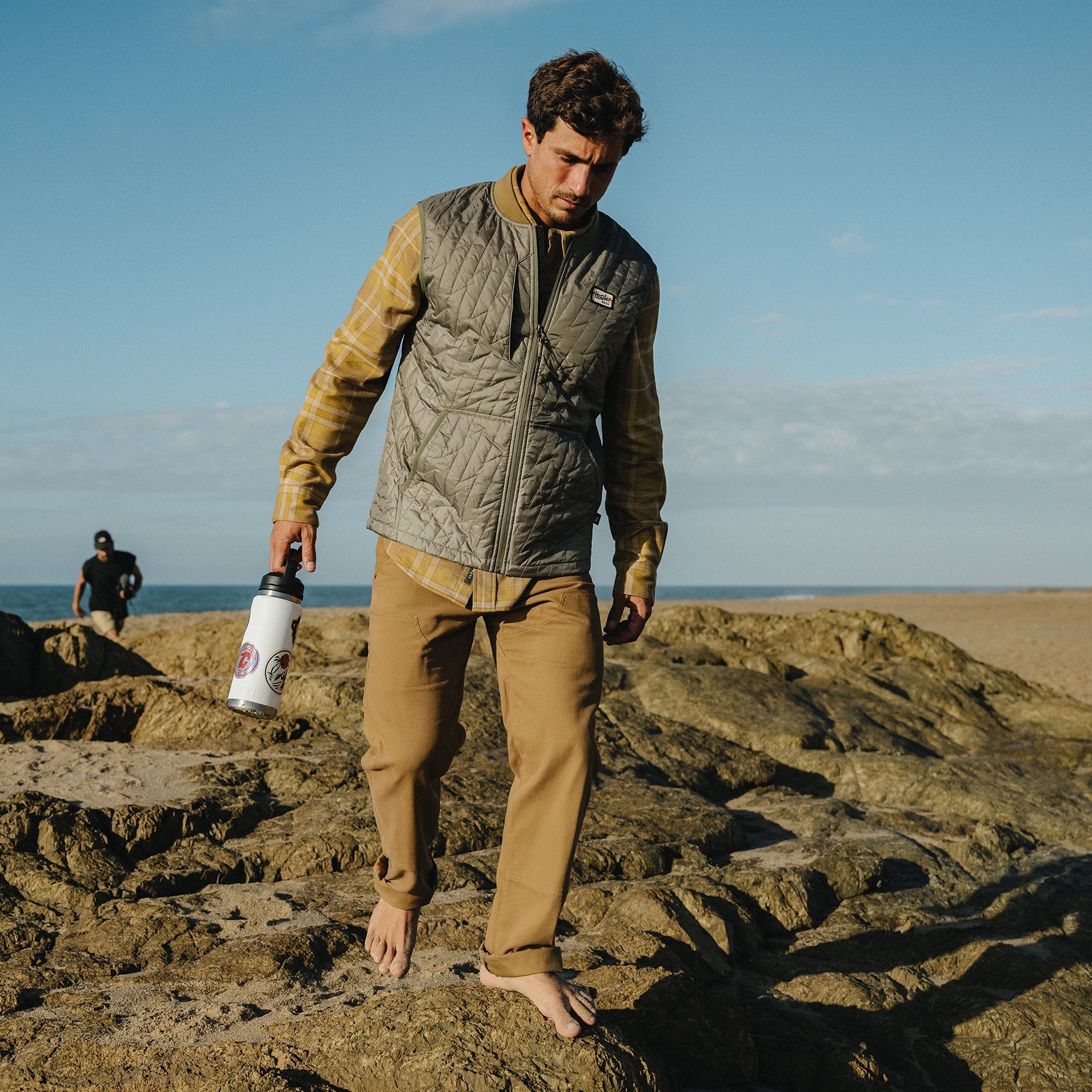 Man walking on rocky beach wearing quilted zip-up vest over plaid flannel shirt, holding water bottle