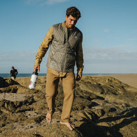 Man walking on rocky beach wearing quilted zip-up vest over plaid flannel shirt, holding water bottle