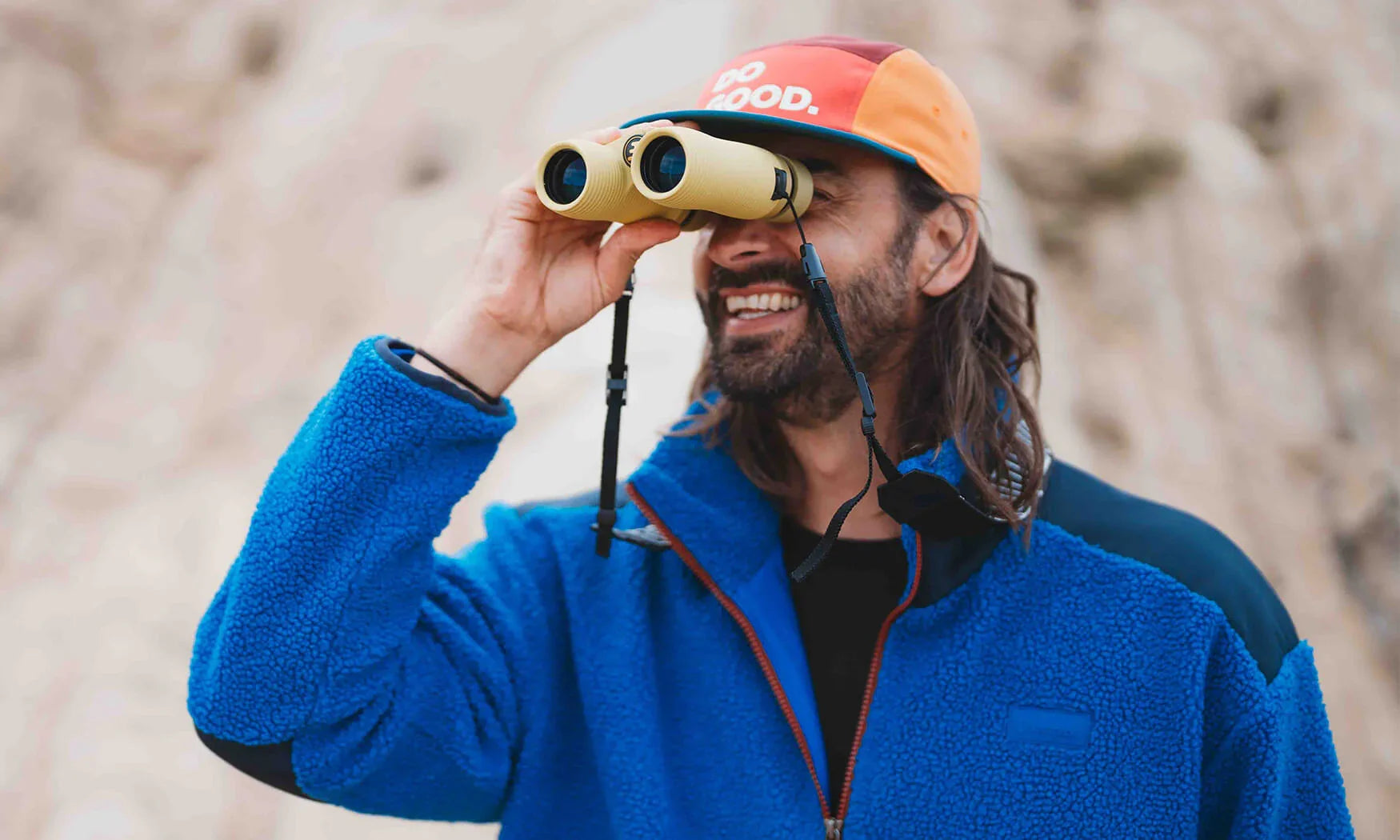Man smiling while looking through compact tan binoculars outdoors, rocky background