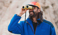 Man smiling while looking through compact tan binoculars outdoors, rocky background