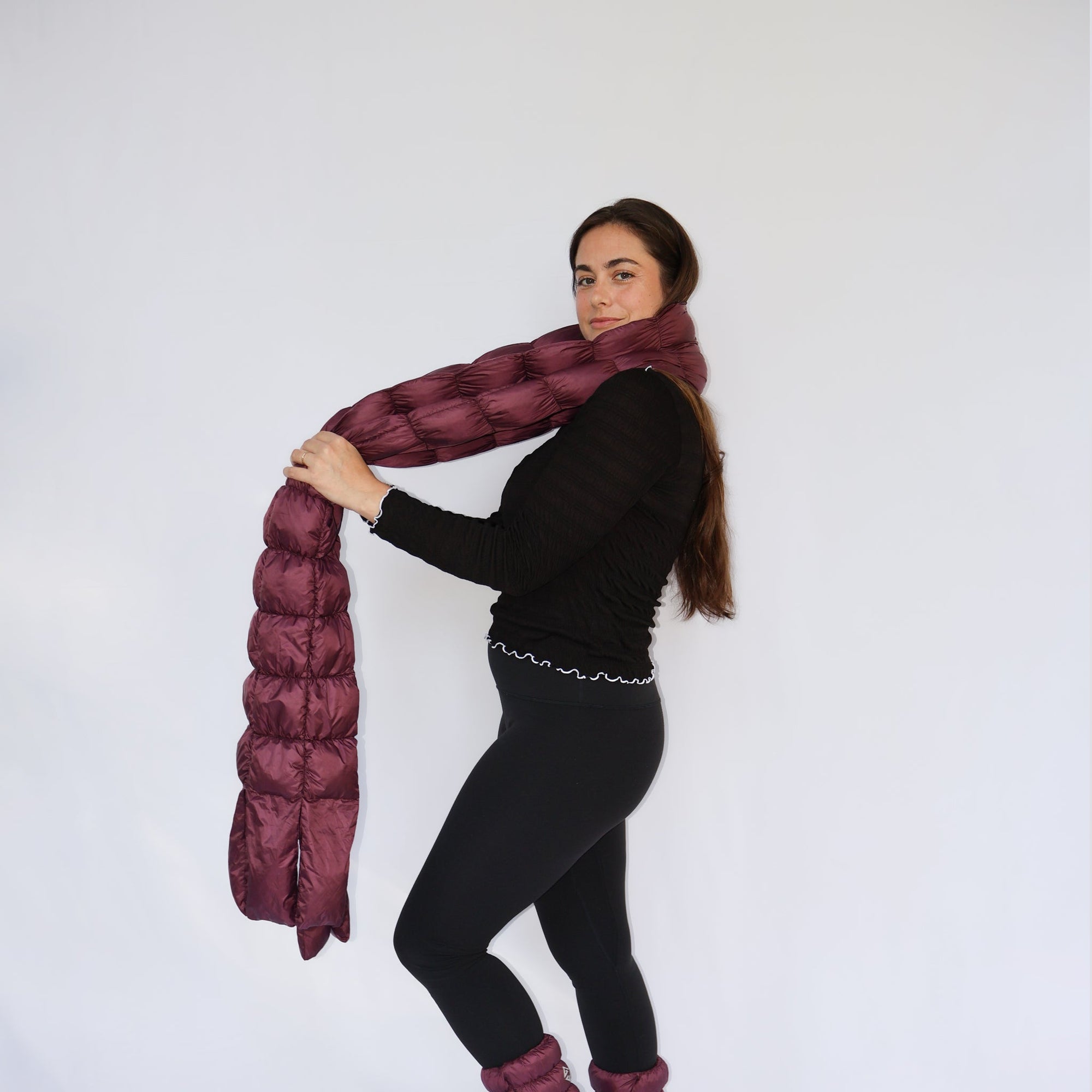 Model wearing a long quilted puffer scarf in deep eggplant, draped around neck with ends held out