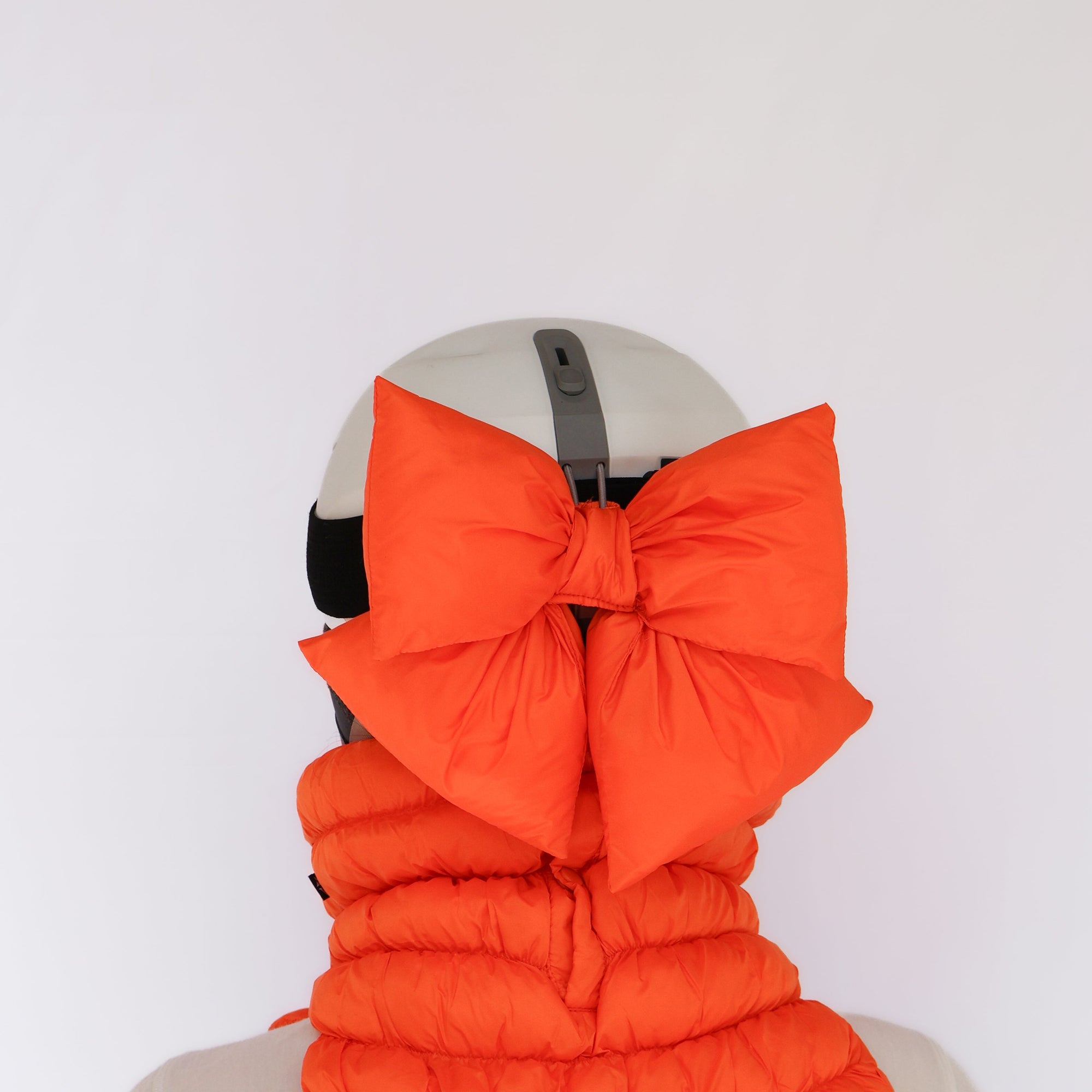 Large puffy bow accessory in bright orange attached to back of white ski helmet