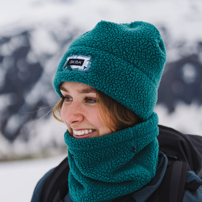 Model wearing a teal high-pile sherpa fleece beanie with brand patch label, snowy mountain background