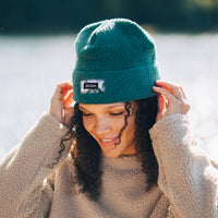 Woman smiling while adjusting a teal high-pile fleece beanie with brand patch on cuff