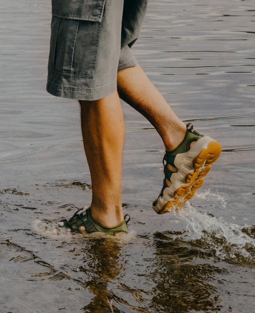 Water shoes with olive green and tan colorway splashing through shallow water on wooden dock