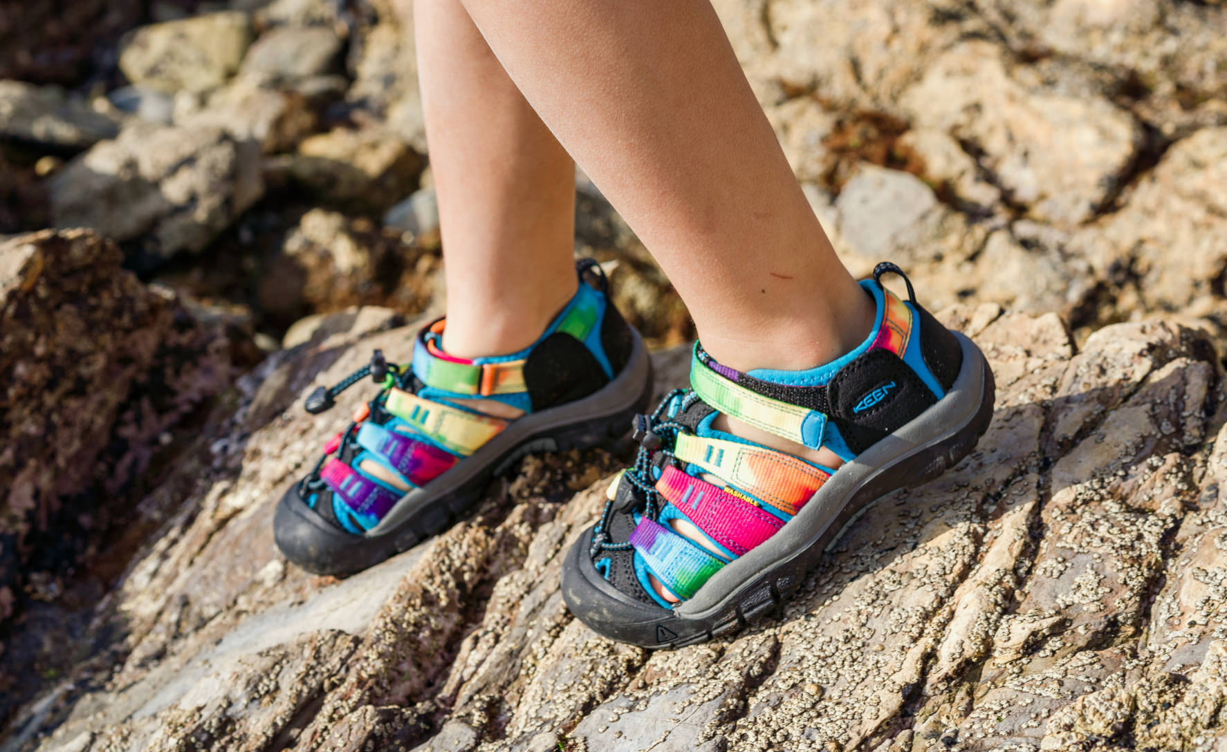 Colorful tie-dye patterned water sandals on child's feet standing on rocky terrain