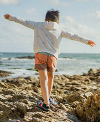 Child wearing colorful sandals standing on rocky coastline with arms outstretched