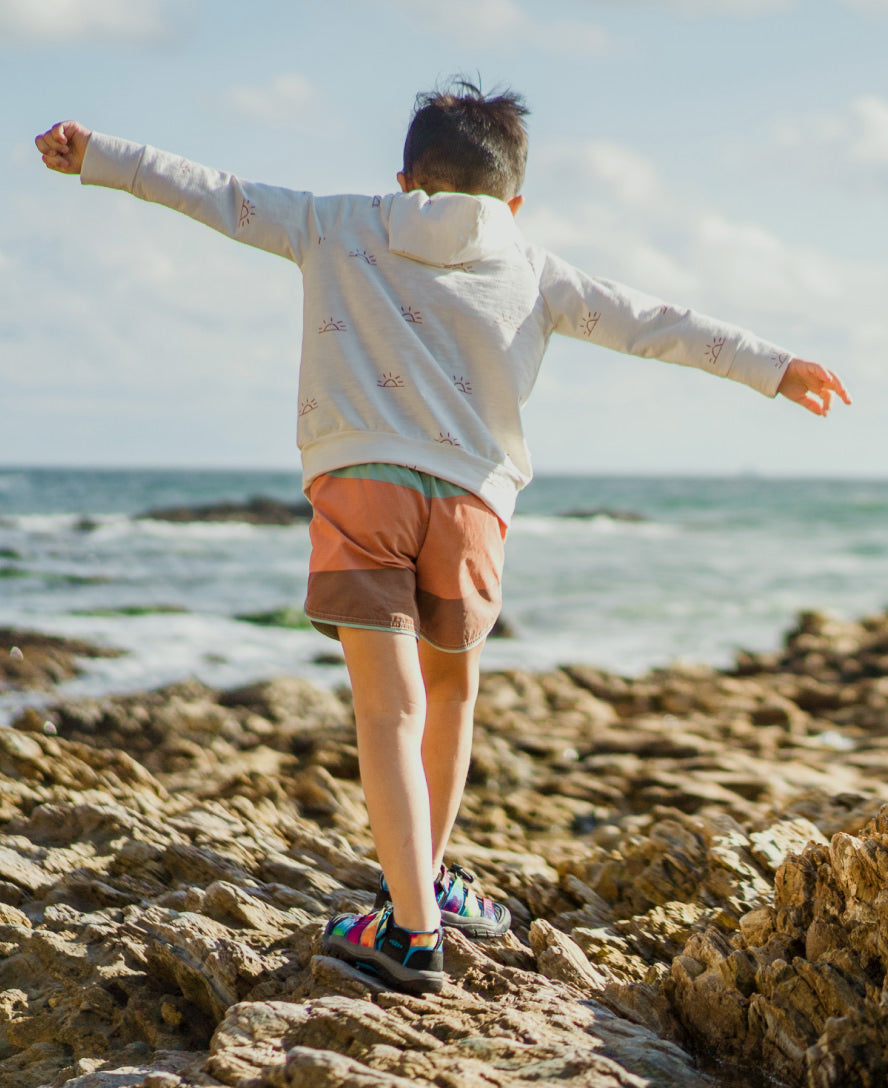 Child wearing colorful sandals standing on rocky coastline with arms outstretched