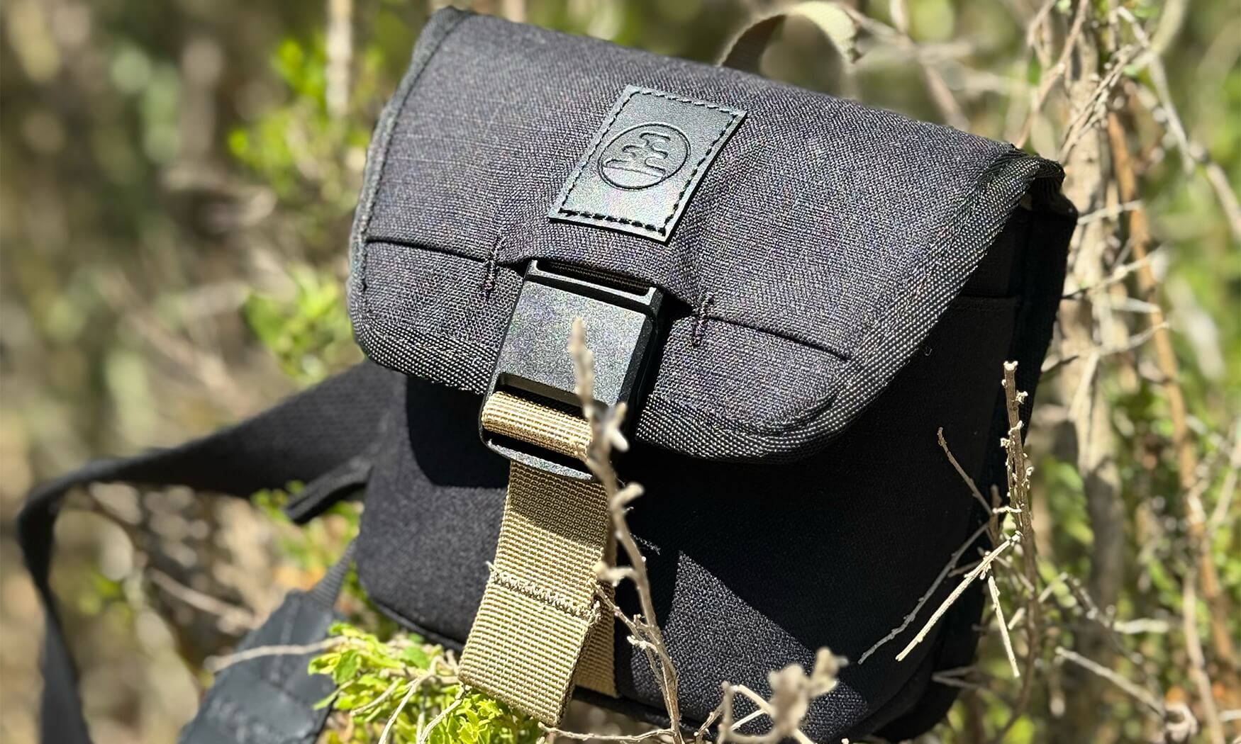 Small dark charcoal field pouch with buckle closure and tan strap, outdoor setting