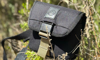 Small dark charcoal field pouch with buckle closure and tan strap, outdoor setting