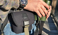 Black canvas belt pouch with buckle closure and tan straps, worn on hip outdoors