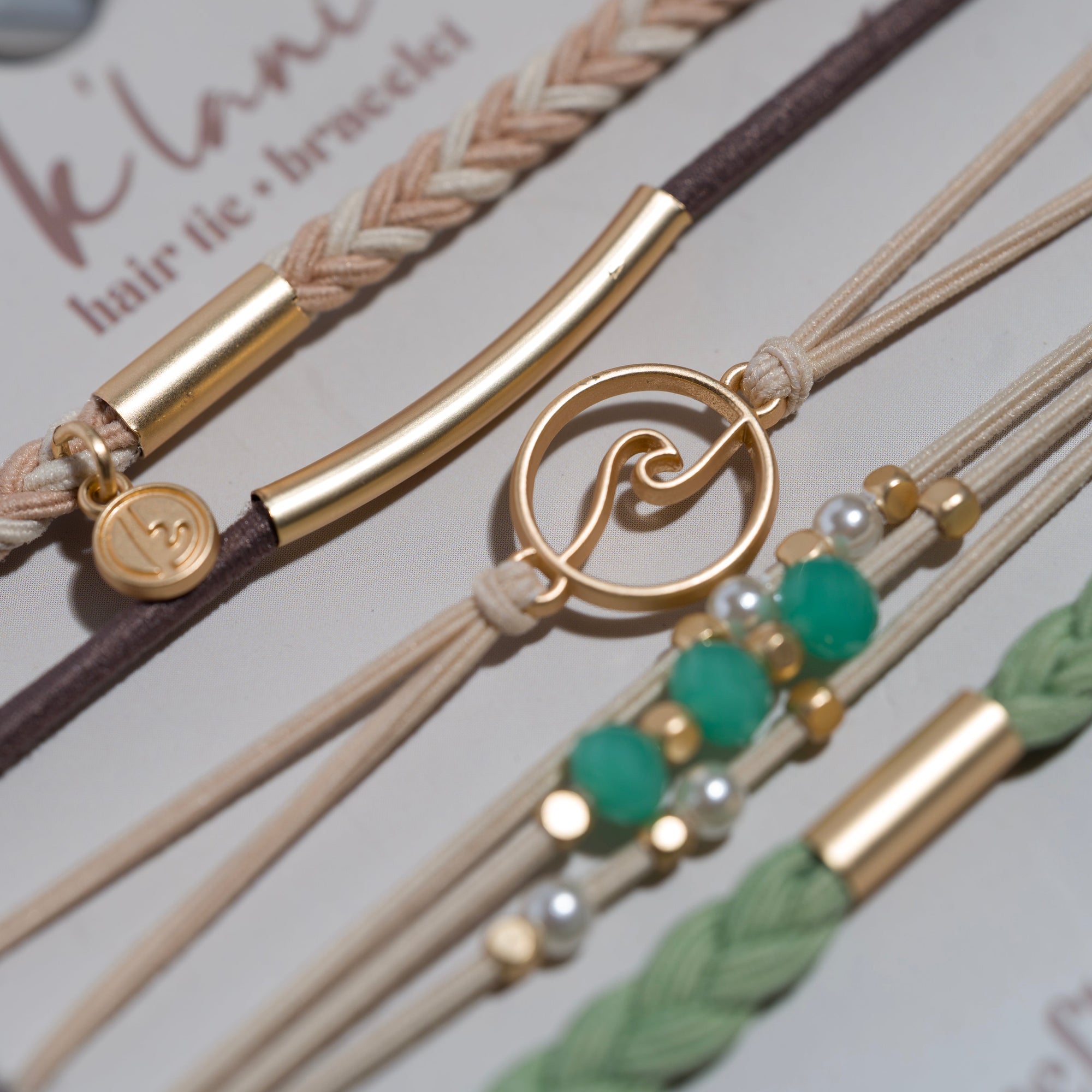 Close-up of multiple hair tie bracelets with gold charms, wave pendant, teal beads, and braided cords