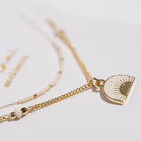 Layered necklace set with gold chain pendant featuring sunburst enamel charm and cream seed bead strand