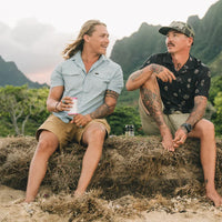 Two men sitting on sandy dunes, one wearing a light blue snap-button short-sleeve shirt with subtle print