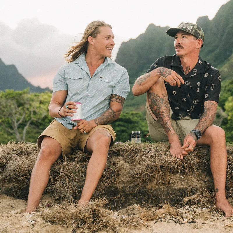 Two men sitting on sandy dunes, one wearing a light blue snap-button short-sleeve shirt with subtle print