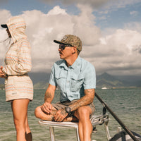 Light blue plaid short-sleeve shirt worn by man sitting on boat with water and mountains