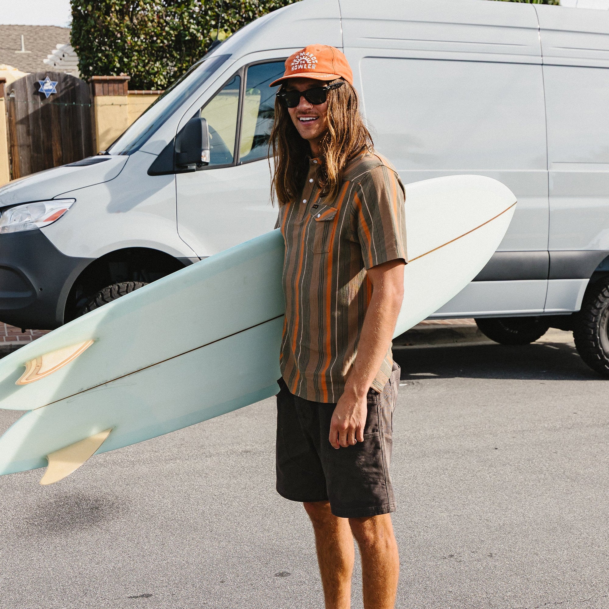 Man carrying surfboard wearing dark brown casual shorts in lifestyle setting