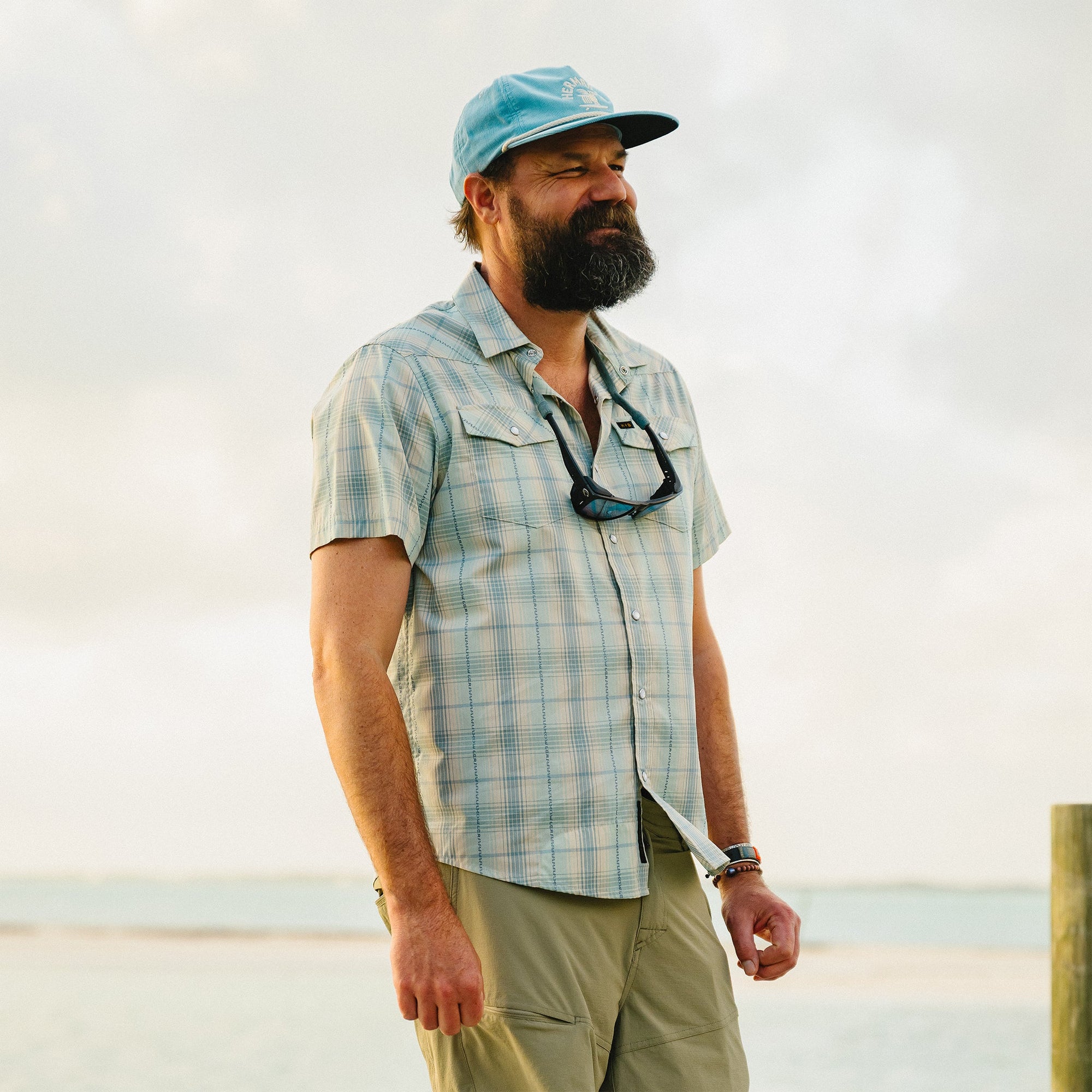 Man wearing plaid short-sleeve snap shirt near ocean, sunglasses clipped to collar, blue cap