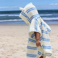 Child wearing white hooded towel with blue stripes on beach, arms outstretched