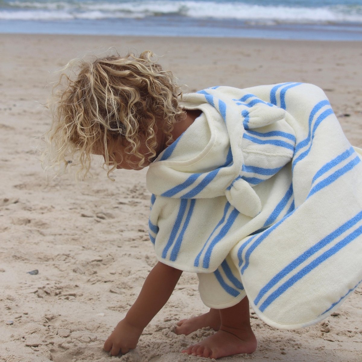 Child wearing white hooded towel with blue stripes on beach sand
