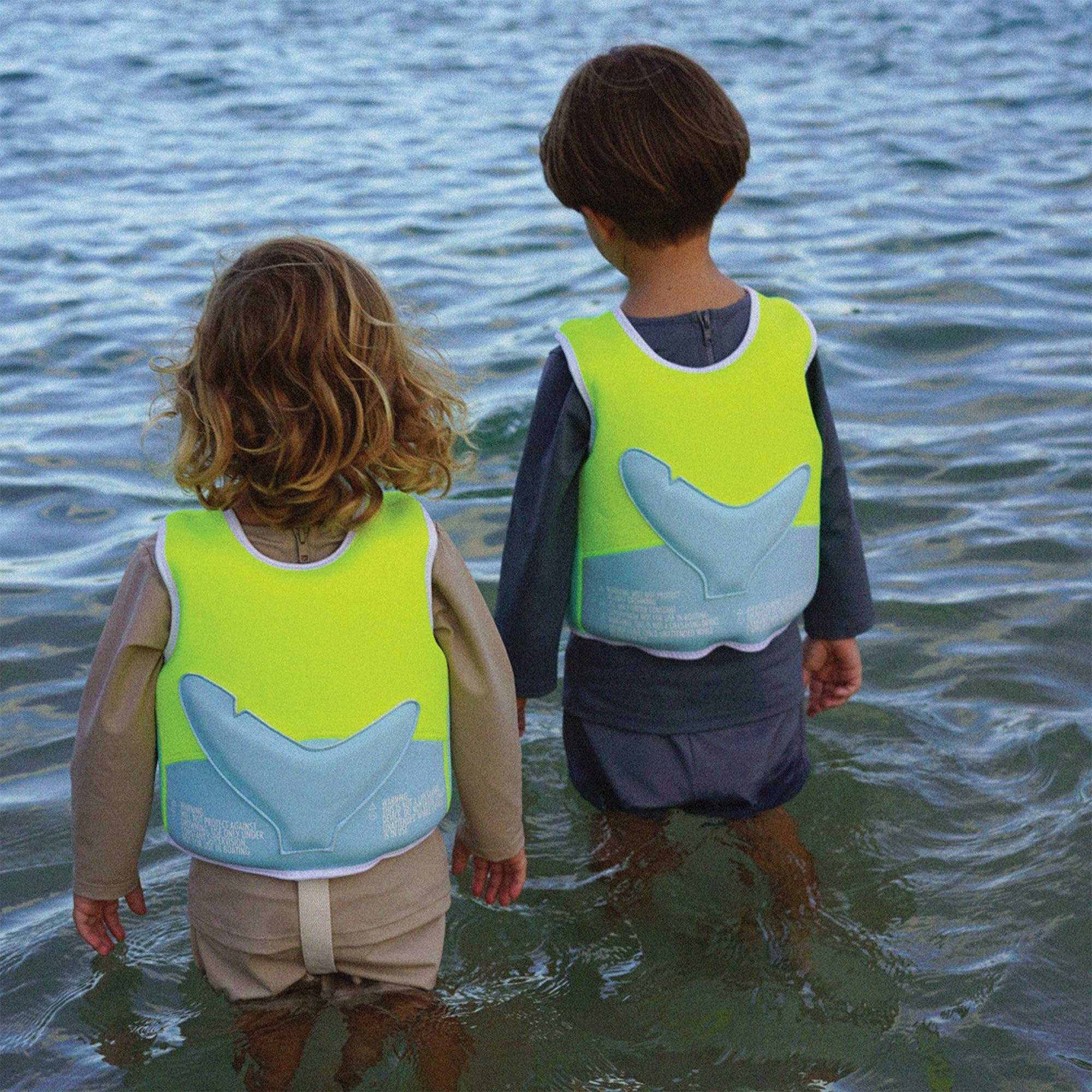 Two children wearing bright green swim vests with whale tail designs, standing in shallow water