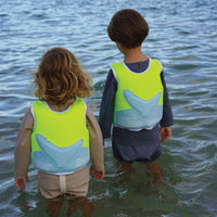 Two children wearing bright green swim vests with whale tail designs, standing in shallow water