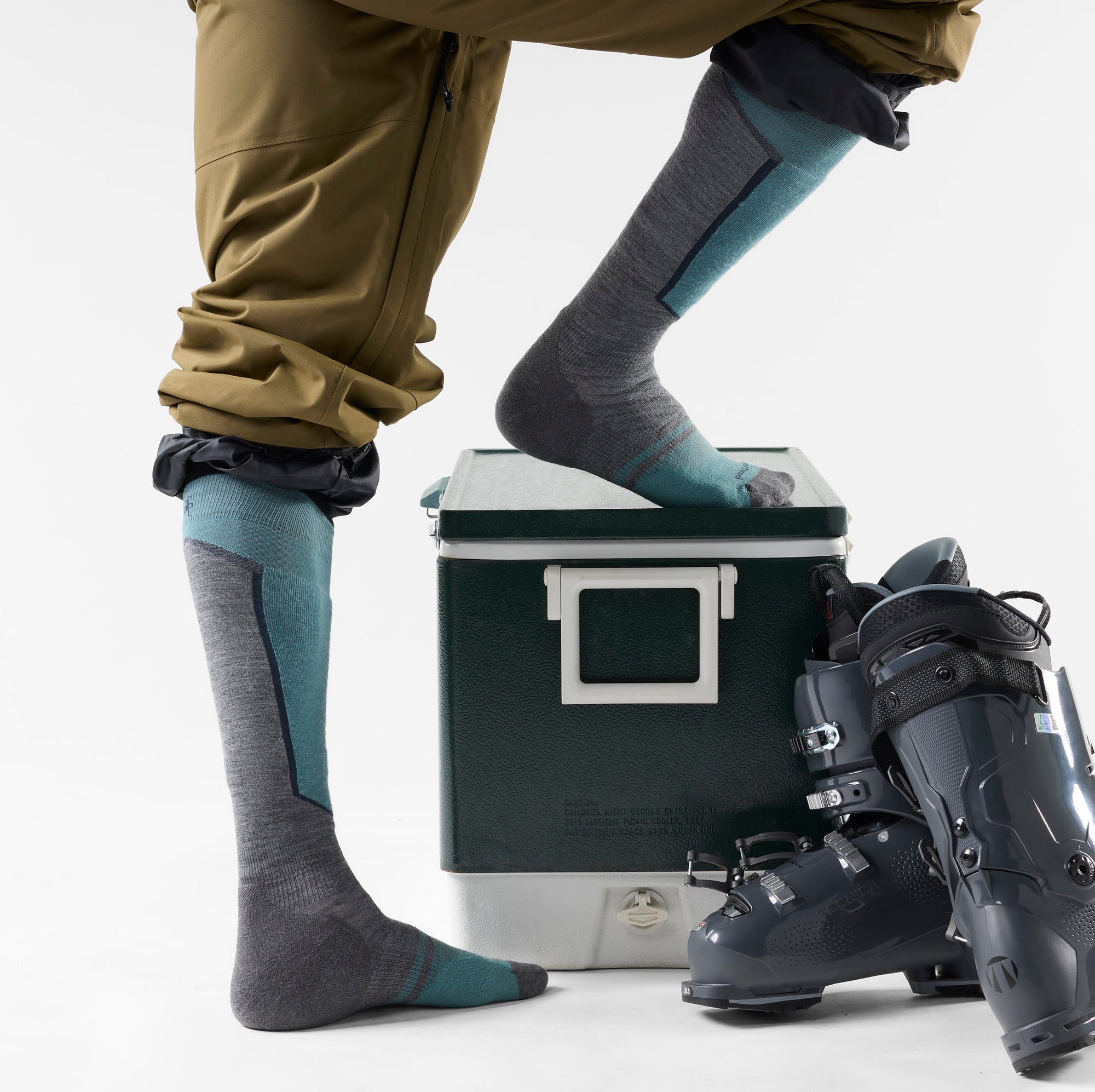 Over-the-calf ski socks in gray with teal accents, shown on model with ski boots nearby