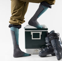 Tall ski socks in gray with teal accents worn on model, one foot propped on cooler, ski boots nearby