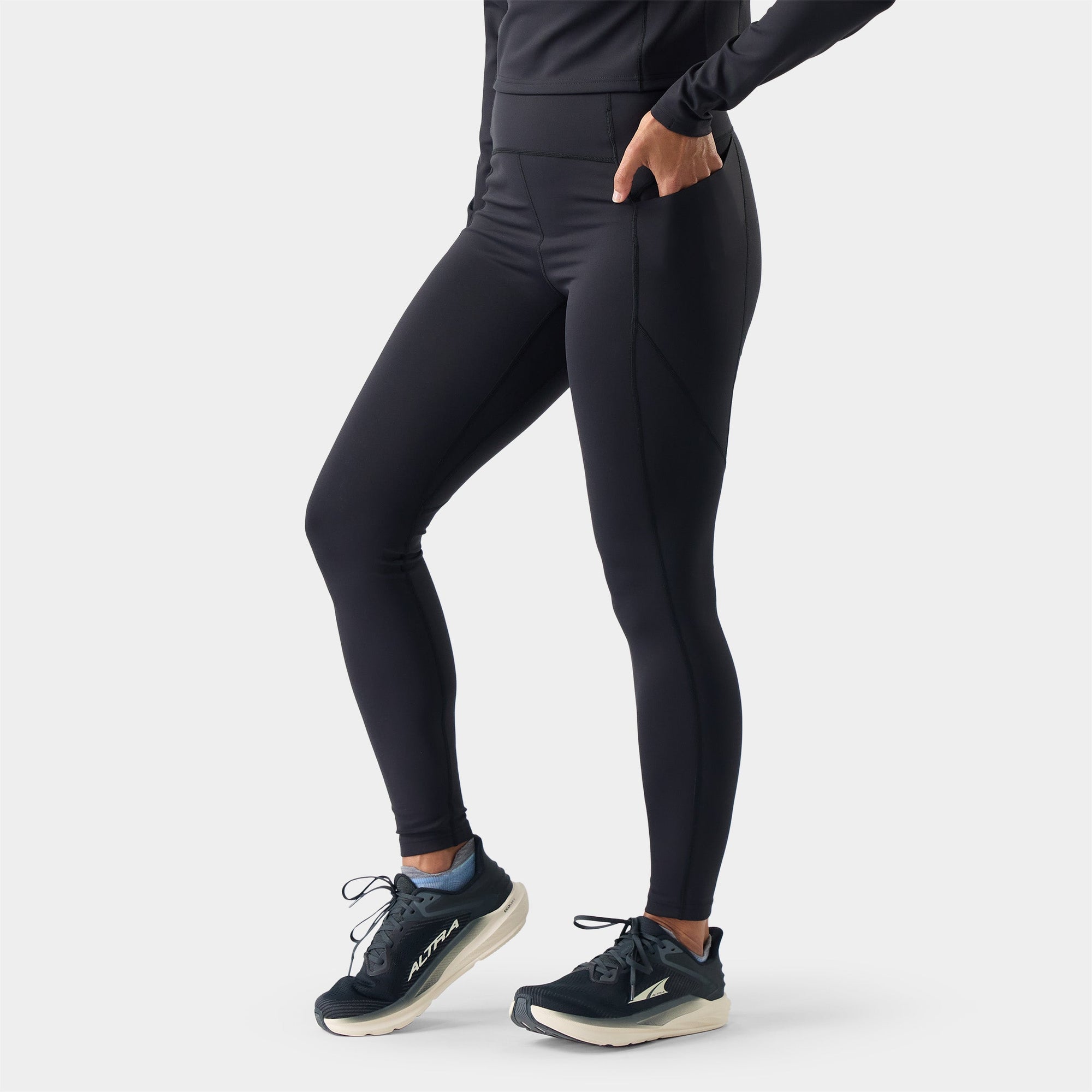 Women's black full-length athletic tights with side pocket, shown from side angle with running shoes