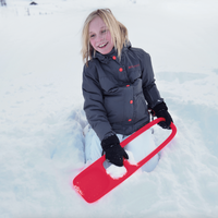 Child holding red snow shovel toy while sitting in snow wearing winter jacket