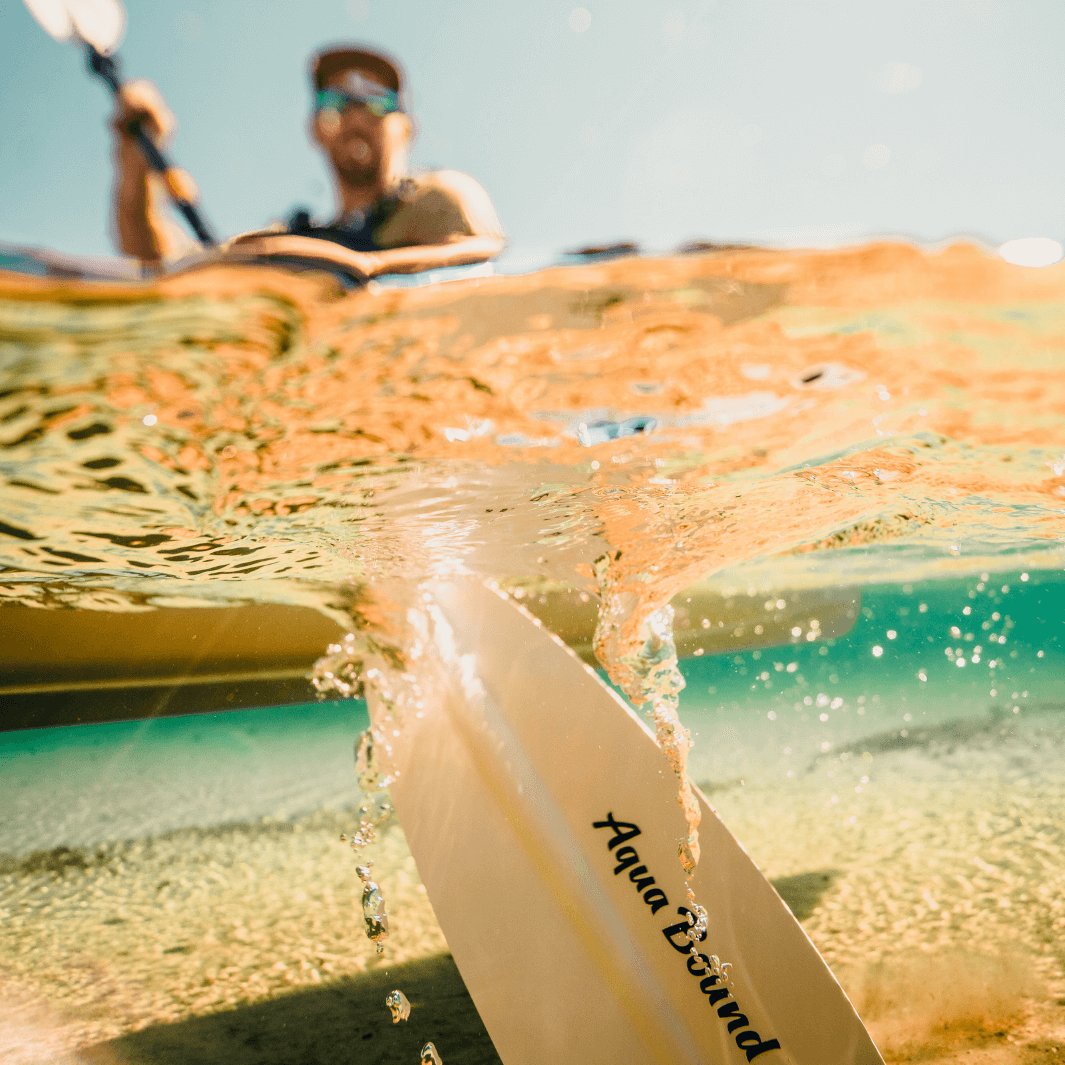 Two-piece kayak paddle blade partially submerged in clear turquoise water with kayaker in background