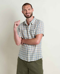 Man wearing light blue plaid short-sleeve button-up shirt with green pants, smiling